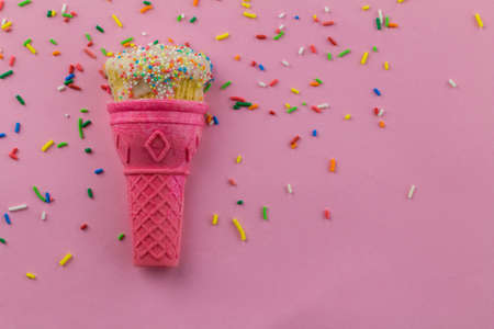 Ice cream cone with little cup cake decorated with frosting and round sprinkles with sprinkles scattered and isolated on pink background - birthday background with copy spaceの写真素材