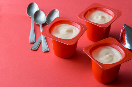 Three red yogurt plastic pots with spoons and foil on red background - creamy fruit yoghurt in plastic cups with copy spaceの写真素材