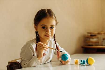 Portrait of a Ukrainian girl. the child paints the egg blue and yellow. The child loves and supports his country. Cute little child girl painting with blue and yellow colors Easter eggs. Hands of a girl with a easter egg. close-up.の写真素材