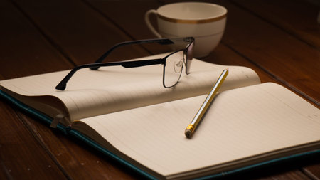 Open notebook with glasses, pencil and cup on wooden desk beside windows.の写真素材