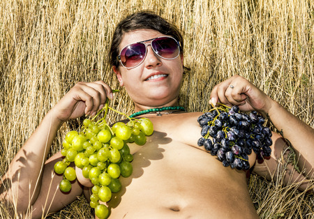 Nude girl posing with bunches of grapes on the chest on the dry grassの写真素材