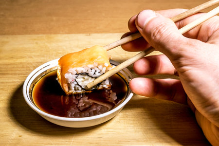 A hand holds sushi sticks and dips them in soy sauce on a wooden surface