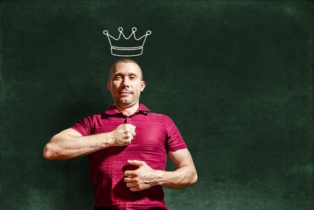 A smug man with a crown on his head hits his chest with his fist against the background of a chalkboardの写真素材