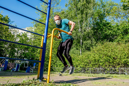 A man in a medical mask trains on a horizontal bar in a courtyard in the city during the pandemic