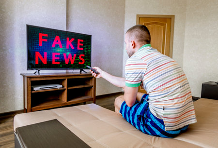 A man is sitting on a sofa and watching TV with fake news on the screen
