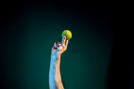 A raised Male hand holds a yellow tennis ball on a dark green background