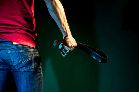 Aggressive man holding a belt in his hand on a dark green backgroundの写真素材
