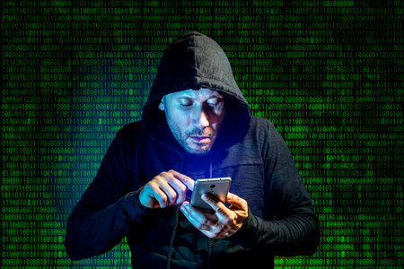 A male intruder with a hood on his head makes a search on a smartphone against the background of...