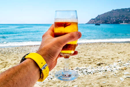 A man holds a glass of cold beer against the hot seascape at the resortの写真素材