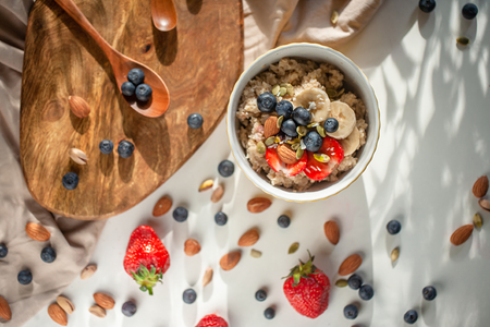 Oatmeal porridge in yellow dish with fruits and nuts on natural wooden tray. Porridge oats with strawberry, pistachios, blueberry, almond and banana. Shadow of sunrise morning. Top view or flat layの写真素材