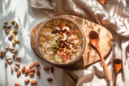 Oatmeal porridge in coconut bowl with wooden spoon on natural wooden tray. Porridge oats with almonds, pistachios and other nuts. Shadow of sunrise morning. White background. Top view or flat lay.の写真素材