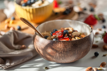 Oatmeal porridge in coconut bowl with wooden spoon. Porridge oats with strawberry, pistachios, blueberry, almond and banana. Shadow of sunrise morning. White background. Side viewの写真素材