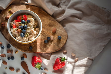 Oatmeal porridge in yellow dish with fruits and nuts on natural wooden tray. Porridge oats with strawberry, pistachios, blueberry, almond and banana. Shadow of sunrise morning. Top view or flat layの写真素材