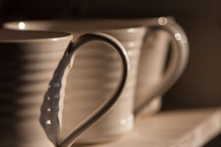 Close-up photo of the handles of white mugs in half-shadow on a shelfの写真素材