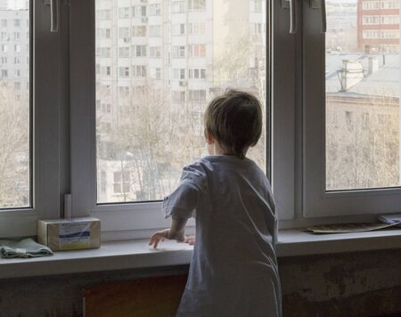 A little boy looks out the window. The street is bright and sunny. Cold weatherの写真素材