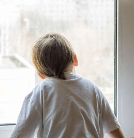 A little boy looks out the window. The street is bright and sunny. Cold weatherの写真素材