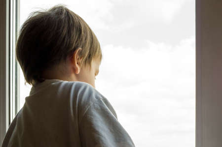 A little boy looks out the window. The street is bright and sunny. Cold weatherの写真素材