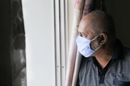 senior man with face mask looking through windowの写真素材