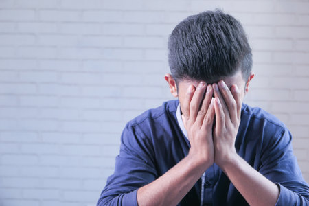 sad man in hood cover face with hands isolated in black.の写真素材
