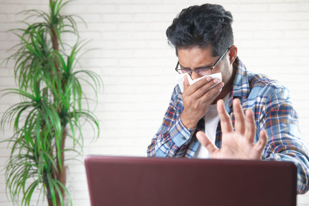 young asian man coughing and sneezes working on laptop