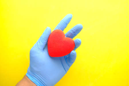 man hand in protective gloves holding red heart on yellow backgroundの写真素材