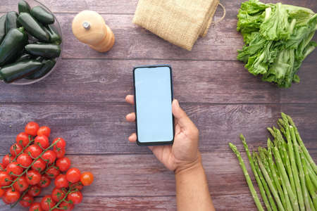 top view of mans hand holding smart phone with fresh vegetable on tableの写真素材