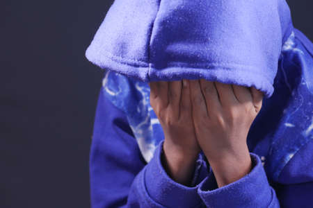 sad man in hood cover face with hands isolated in black.の写真素材