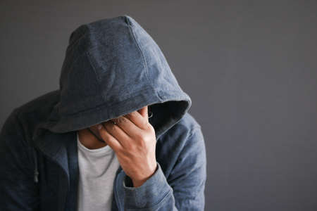 sad man in hood cover face with hands isolated in black.の写真素材