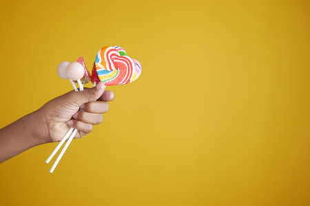 child holding rainbow heart shape lollipop against yellow backgroundの写真素材