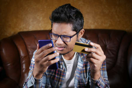 upset man holding credit card and smart phone while sited on sofaの写真素材