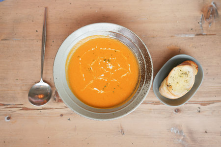 top view of a bowl of yellow tomato vegetables soup and garlic bread on table