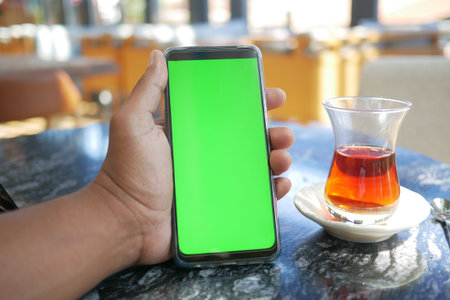 holding smart phone with green screen and tea on table