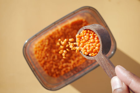 A hand holds a measuring spoon of red lentils above a container ready to be filled