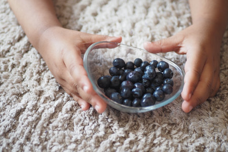 A bowl of fresh blueberries in youthful hands showcases the essence of childhood snacksの写真素材