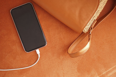 A modern smartphone is conveniently being charged next to a stylish handbag on a soft surfaceの写真素材
