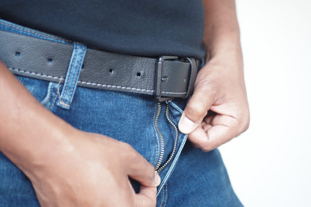 Individual adjusting zipper on blue jeans while wearing a black belt indoors