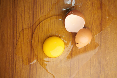 Egg yolk spills next to broken eggshells on wooden surface