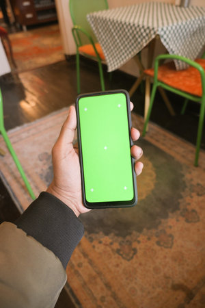 Hand holding a smartphone with a blank green screen in a cafe