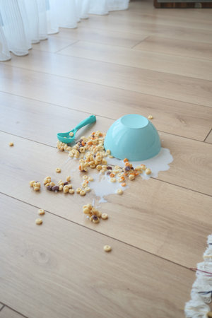 A blue bowl has tipped over, spilling cereal and milk on the floor
