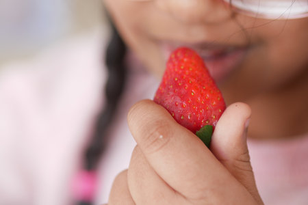 Little girl enjoying a fresh strawberry in bright sunlightの写真素材