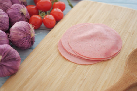 Sliced deli meat on a wooden board with fresh vegetables nearbyの写真素材