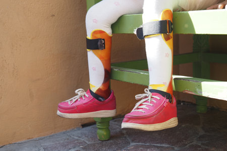 Child with colorful leg braces sitting on a green wooden benchの写真素材