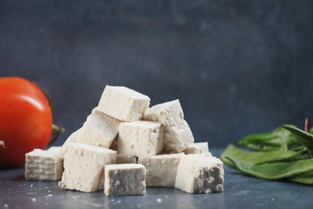 Fresh tofu cubes arranged with tomato and greens on a dark surfaceの写真素材