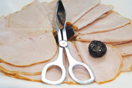Sliced deli meat arranged with scissors and black olives on a plate
