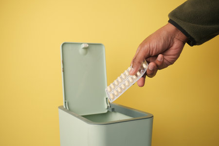 Disposing of medication in a green trash bin against yellow backgroundの写真素材