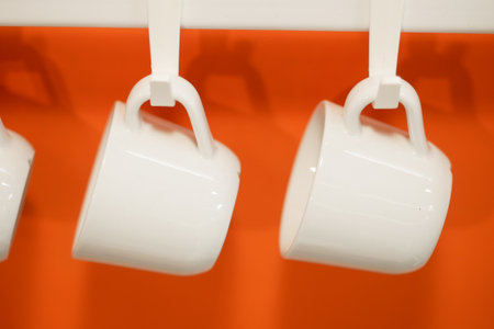 White ceramic mugs hanging on a bright orange wall in a cafeの写真素材