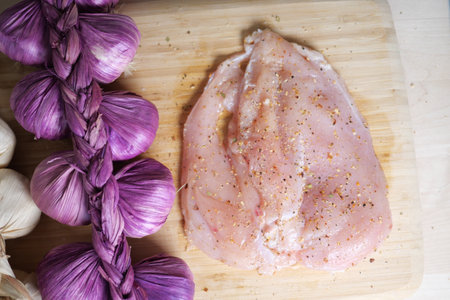 Chicken breast seasoned and ready for cooking next to garlic and onionsの写真素材