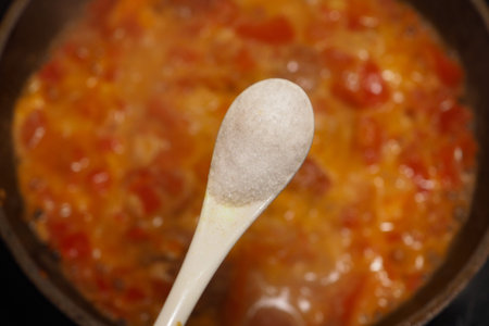 A spoon with salt is above bubbling tomato sauce on the stove