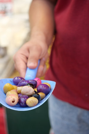 A hand holds a blue scoop filled with colorful olives at a local marketの写真素材