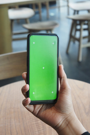 Hand holding smartphone with green screen in cafe setting